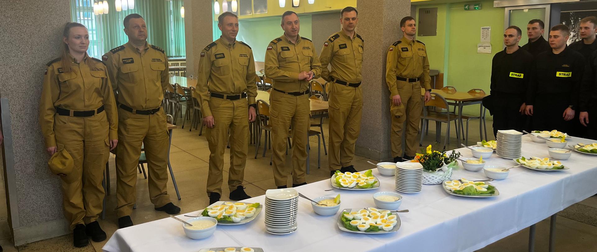 Wnętrze stołówki SA PSP w Krakowie. Nakryte białymi obrusami stoły, na nich białe talerze i tace z przekrojonymi na pół jajkami. Dookoła stu strażacy. Ludzie w umundurowaniu służbowym i koszarowym. 