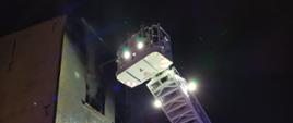 Strażacy w koszu drabiny mechanicznej gaszą pożar mieszkania w budynku mieszkalnym wielorodzinnym na wysokości drugiego piętra.