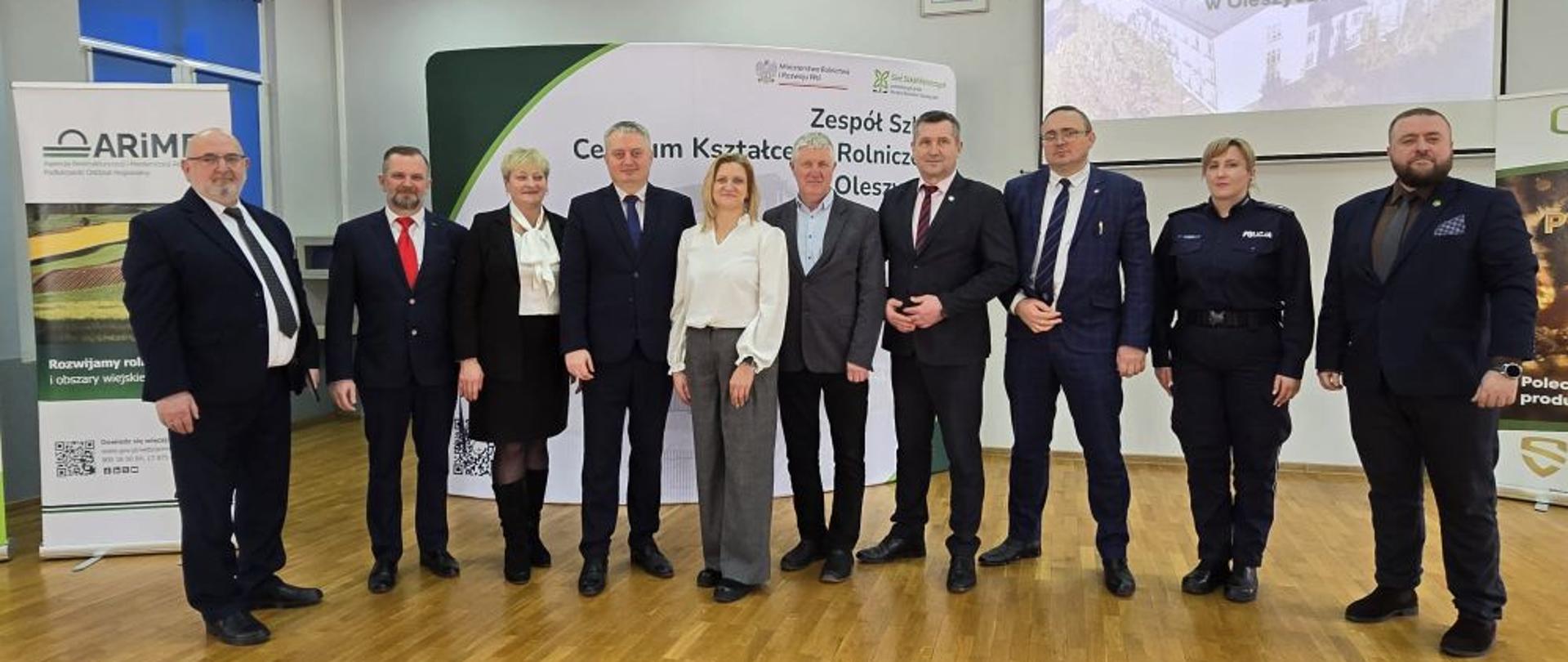 Prelegenci i zaproszeni goście stoją na scenie, w tle banery reklamowe i prezentacja multimedialna