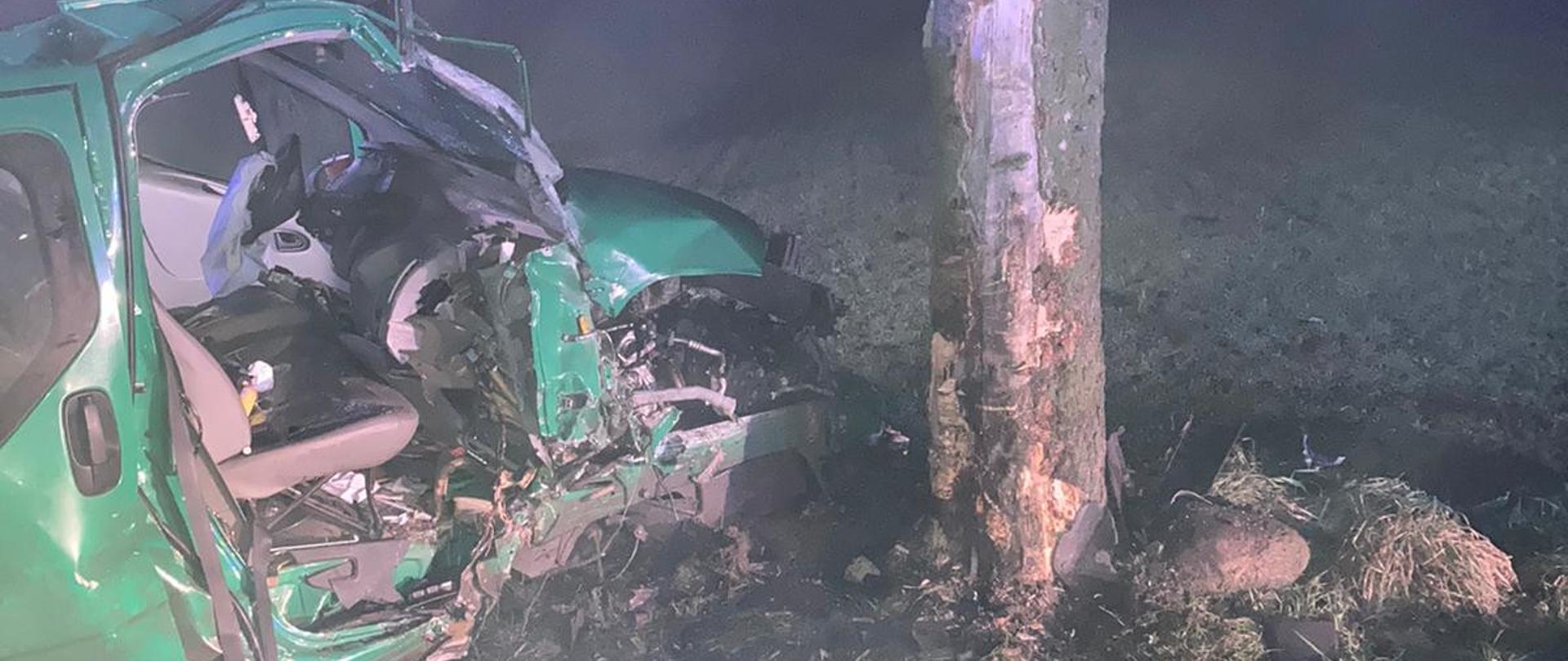 Na zdjęciu miejsce wypadku po uderzeniu w przydrożne drzewo. Widoczna jest uszkodzona prawa strona pojazdu dostawczego Opel VIVARO koloru zielonego. Widoczne jest również drzewo po uderzeniu z pojazdem, gdzie została uszkodzona kora drzewa. 