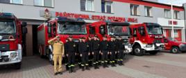Strażacy w dwuszeregu na tle samochodów strażackich.