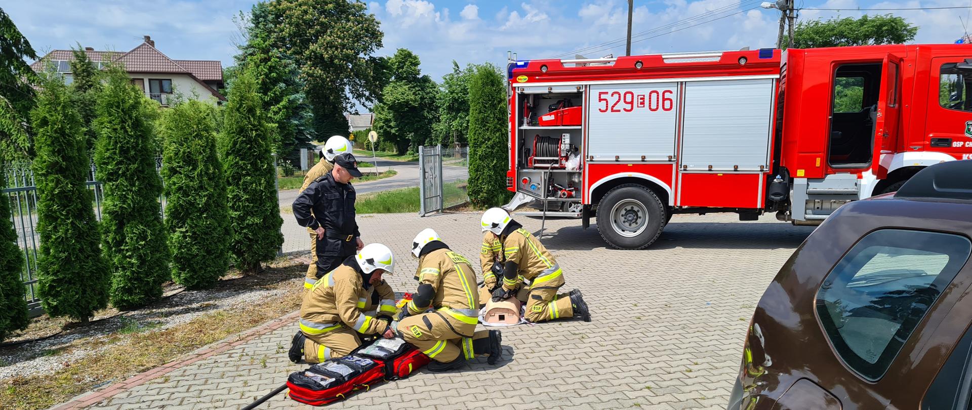 Zdjęcie przedstawia 4 strażaków udzielających pierwszej pomocy fantomowi, w ramach ćwiczeń, a obok nich znajdują się 2 strażacy stojący nad nimi i obserwujący ich pracę. Wszyscy ubrani są w stroje strażackie bojowe i znajdują się na podjeździe/parkingu remizy. Za nimi stoi samochód strażacki.