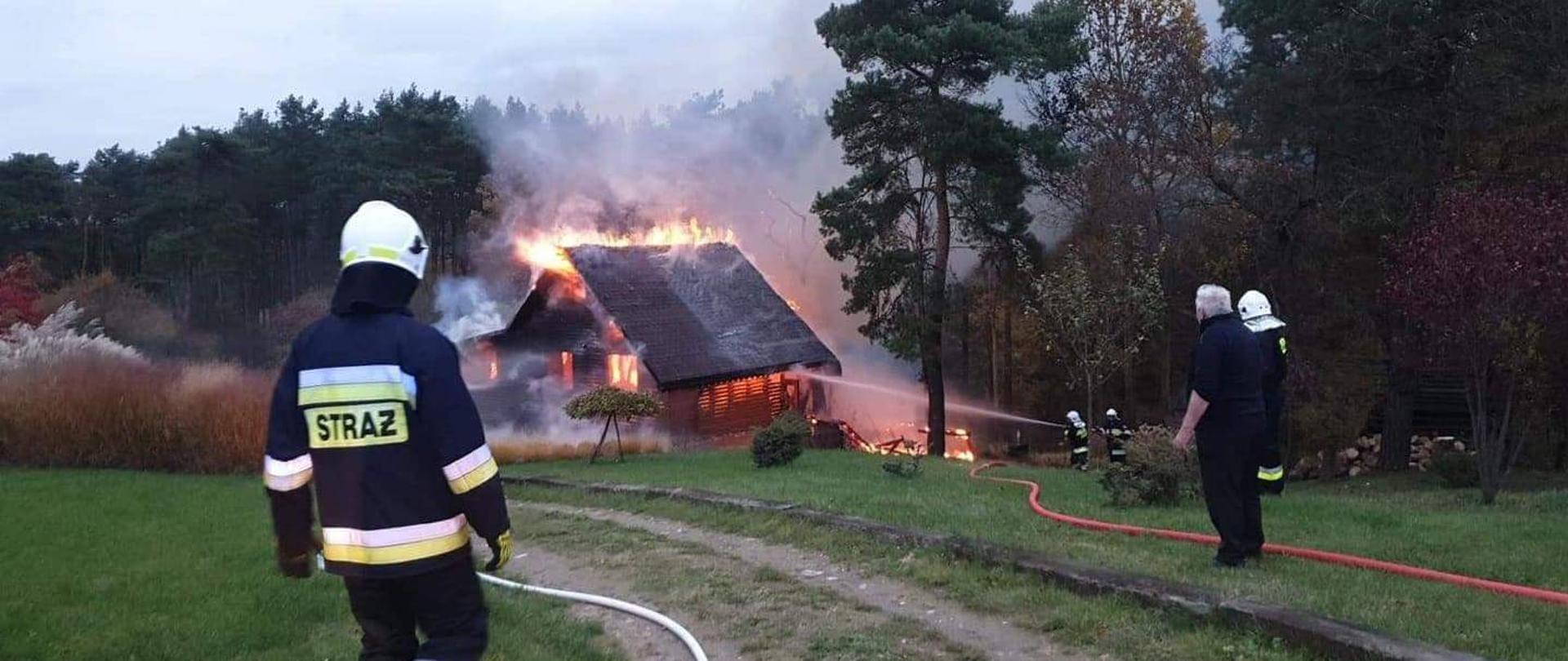Na zdjęciu widzimy palący się dom oraz strażaków walczących z pożarem 