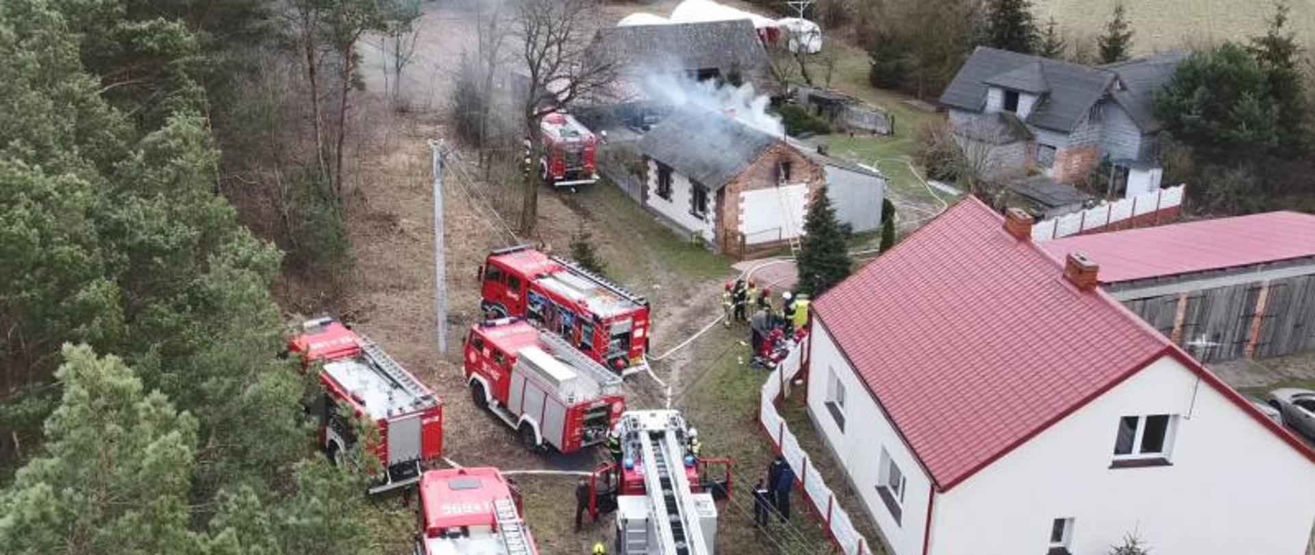 Zdjęcie przedstawia widok pożaru budynku mieszkalnego z lotu ptaka wokół, którego znajduje się sześć samochodów bojowych pożarniczych oraz strażaków udzielających pomocy dwóm osobom poszkodowanym- poparzonym w wyniku pożaru. Po bokach zdjęcia znajdują się inne budynki mieszkalne oraz las.
