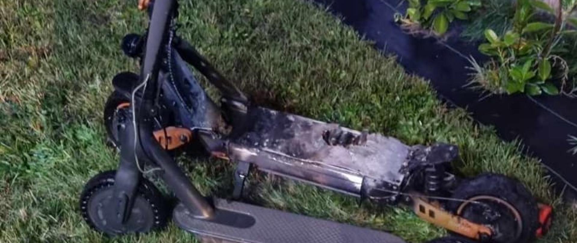 Zdjęcie przedstawia uszkodzoną hulajnogę elektryczną przez spalony akumulator. Hulajnoga stoi na trawniku przy betonowym płocie. 