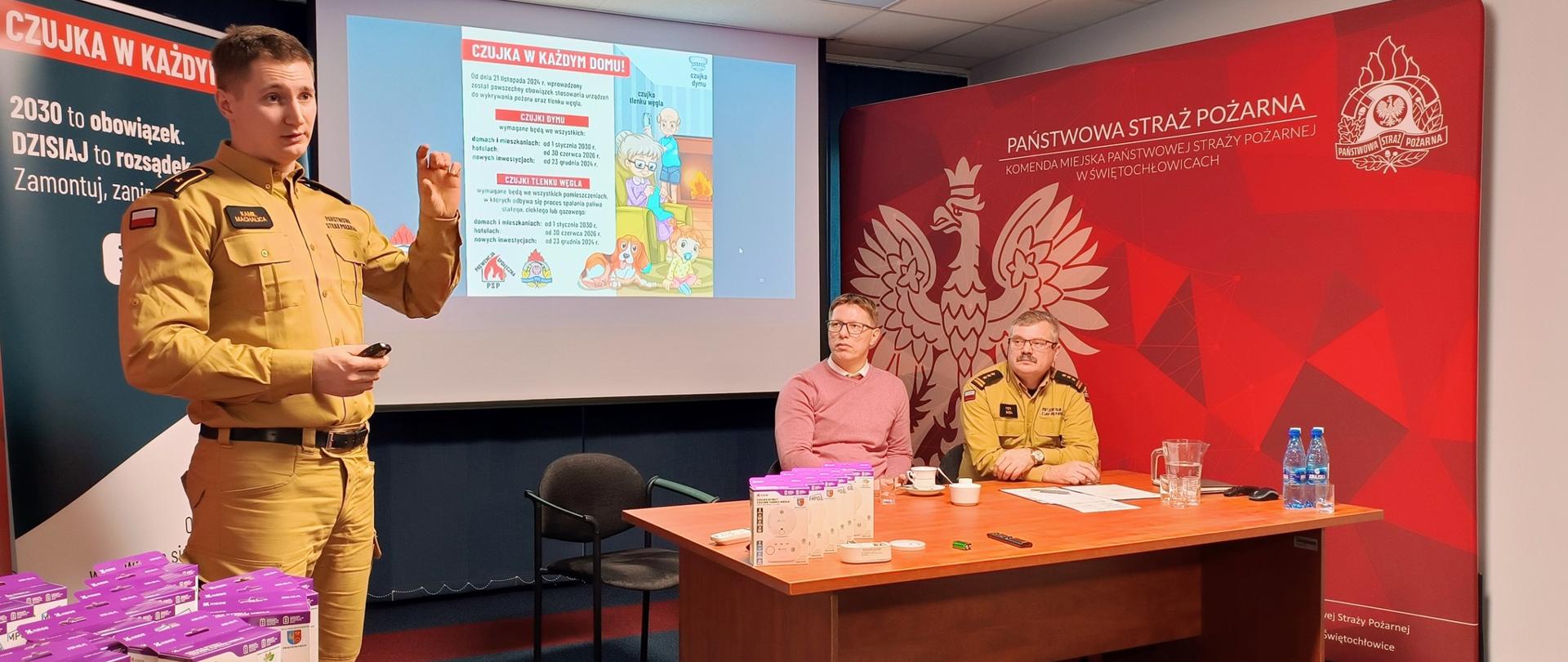Zdjęcie przedstawia kluczowy moment konferencji poświęconej kampanii "Czujka w Każdym Domu". Na pierwszym planie, po lewej stronie, stoi funkcjonariusz Państwowej Straży Pożarnej w piaskowym mundurze, dynamicznie przemawiający do publiczności i prezentujący mały przedmiot. Obok niego, na małym okrągłym stoliku, widoczne są stosy białych opakowań czujników i czujek. W tle, na dużym ekranie, wyświetlany jest slajd informacyjny z hasłem kampanii, co podkreśla jej edukacyjny charakter i znaczenie.
Za dużym, drewnianym biurkiem siedzą dwie osoby – Prezydent w łososiowym swetrze oraz drugi oficer PSP w piaskowym mundurze. Obaj obserwują prezentującego. Tło spotkania stanowi duża, czerwona ściana z wizerunkiem godła państwowego oraz pełnym napisem "PAŃSTWOWA STRAŻ POŻARNA KOMENDA MIEJSKA PSP W ŚWIĘTOCHŁOWICACH", co świadczy o oficjalnym i lokalnym charakterze wydarzenia. Scena ta silnie koncentruje się na promowaniu bezpieczeństwa i gotowości służb do wspierania inicjatywy prewencyjnej.
