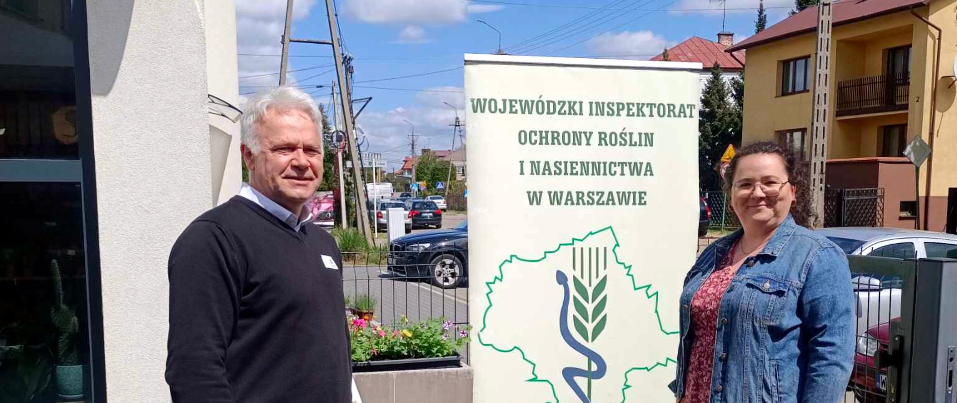 Na zdjęciu dwoje osób stoi na zewnątrz obok stolika i roll-upu z logo oraz nazwą Wojewódzkiego Inspektoratu Ochrony Roślin i Nasiennictwa w Warszawie. Na stoliku znajdują się materiały promocyjne. W tle widoczne są budynki mieszkalne i ulica.