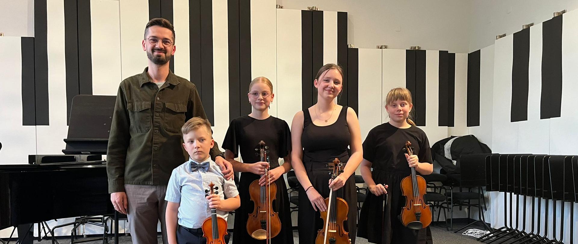 Nauczyciel stoi na scenie z uczniami, którzy trzymają skrzypce.