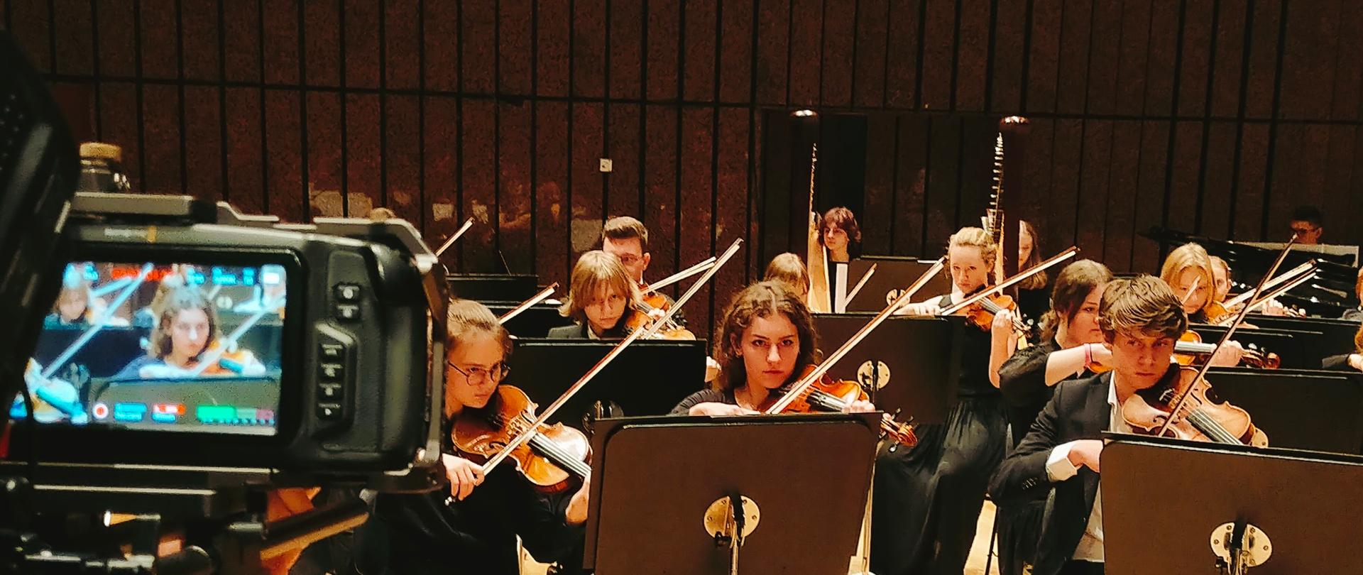 wielu uczniów w zespole orkiestry gra koncert na scenie. Po lewej widoczna kamera która ich nagrywa.
