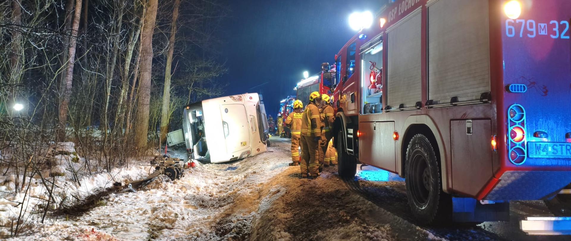 Tył przewróconego w rowie autobusu. Po prawej stronie samochód pożarniczy i strażacy.