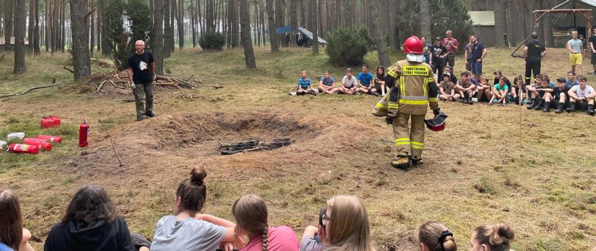 Zdjęcie przedstawia grupę młodzieży siedzącą po okręgu wokół strażaka prowadzącego pogadankę. Strażak ma na sobie ubranie specjalne oraz hełm. Przed nim widać gaśnice, które będą wykorzystane do pokazów gaszenia ognia w specjalnie przygotowanym miejscu. W oddali widać drzewa oraz namioty.