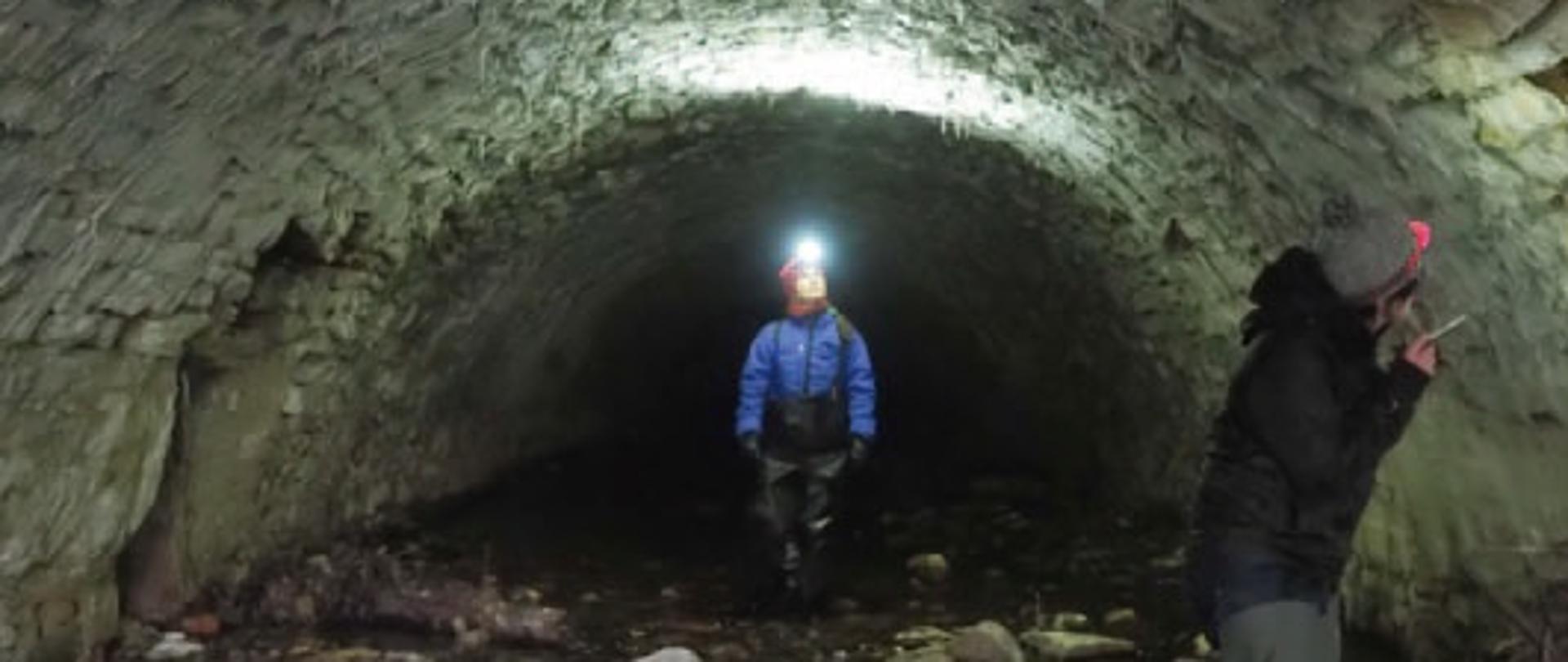 Dwie postacie z latarkami czołówkami przyglądają się ścianom i szczelinom w półkolistym, kamiennym tunelu.