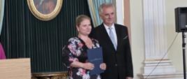 na zdjęciu na tle ciemnej ściany ze obrazem w złotej ramie kobieta i meżczyna elegancko ubrani kobieta trzyma w rękach niebieską teczkę