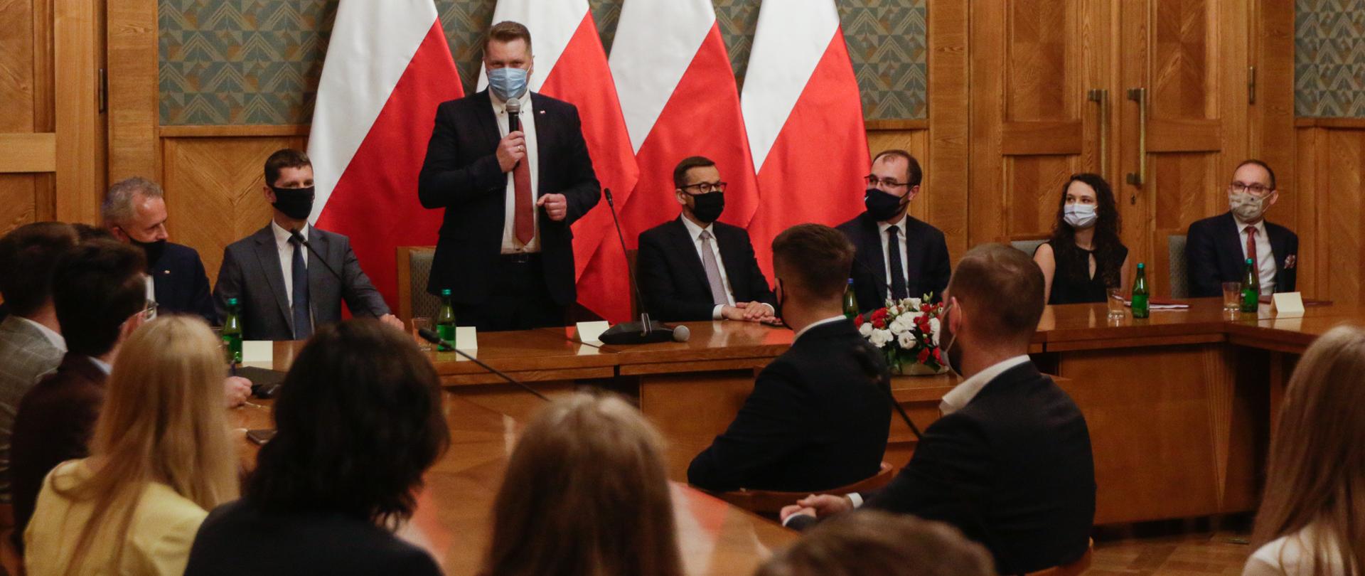 Minister Czarnek przemawia podczas spotkania. Obok siedzą premier Mateusz Morawiecki oraz wiceminister Dariusz Piontkowski. Na przeciwko siedzą pozostali uczestnicy spotkania.