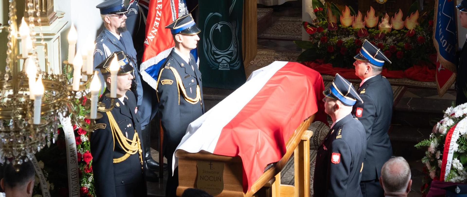 W kościele trwa uroczystość pogrzebowa; trumna okryta biało-czerwoną flagą otoczona jest przez strażaków w wyjściowych czarnych mundurach ze złotymi obszyciami oraz funkcjonariuszy służby więziennej.