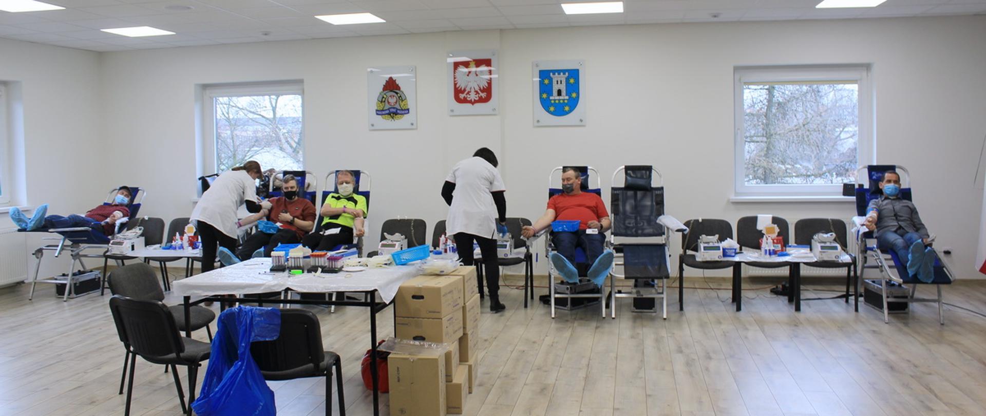 Zdjęcie sali, na której rozstawione są stanowiska do oddania krwi. Na pierwszym planie widać stolik, na których są niezbędne rzeczy pielęgniarek. Tuż za stolikiem w szeregu rozstawione są naprzemiennie krzesła i specjalne fotele do oddania krwi, na których siedzą mężczyźni oddający krew. Obok Panów są pielęgniarki, które nadzorują cały proces. 