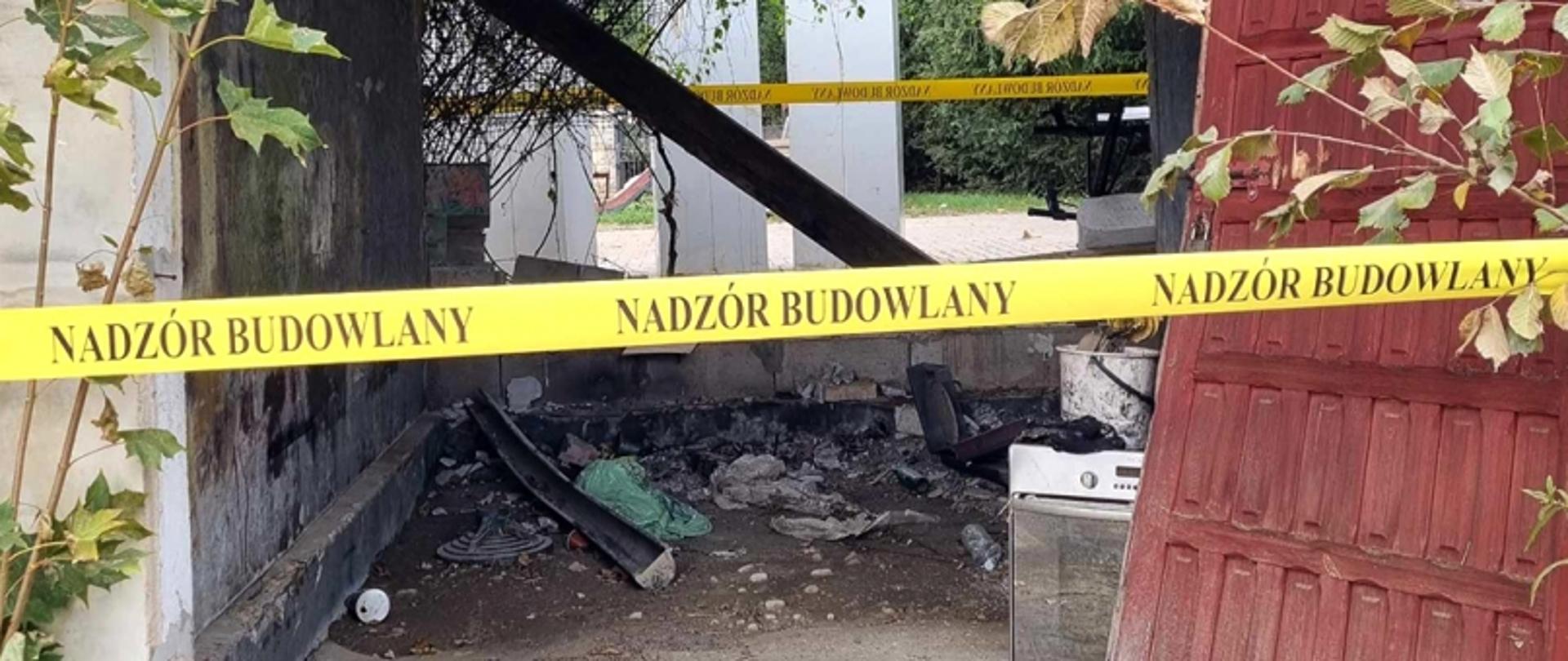 Zniszczona konstrukcja z taśmą ostrzegawczą, gruzem i częściami urządzeń; wokół roślinność i drewniane podpory.