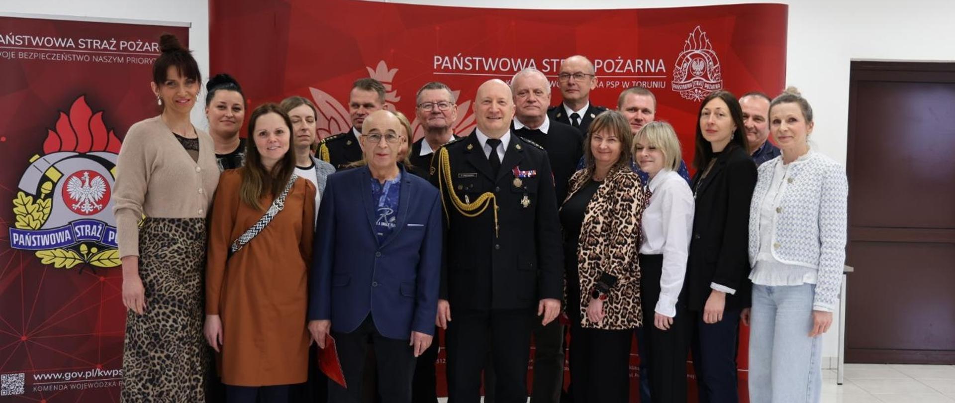 Zdjęcie zbiorowe kilkunastu osób, gdzie są mundurowi , a w większości cywile, kobiety i mężczyźni. Za nimi ścianka firmowa w kolorze czerwonym.