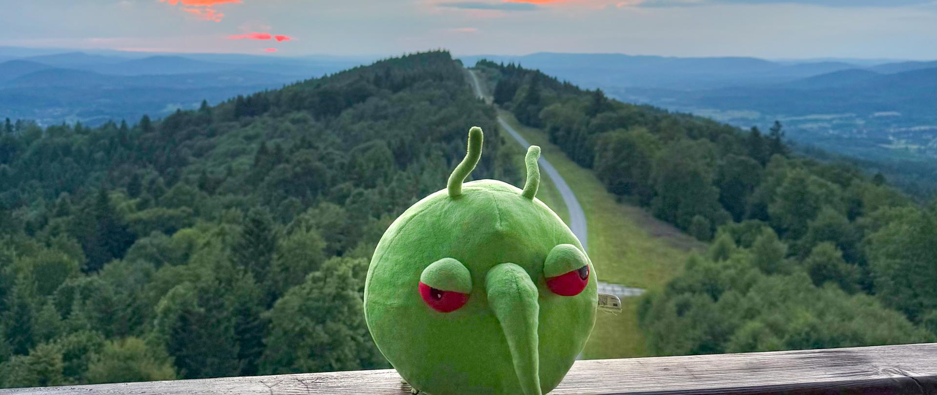 Zielona okrągła maskotka siedząca na balustradzie drewnianej. Z tylu widok na góry i niebo z pomarańczowo - czerwoną barwą zachodzącego słońca.