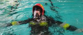 Strażak w hełmie, ubraniu specjalnym i aparacie ochrony układu oddechowego pływa w basenie. W tle widać innych strażaków w wodzie.