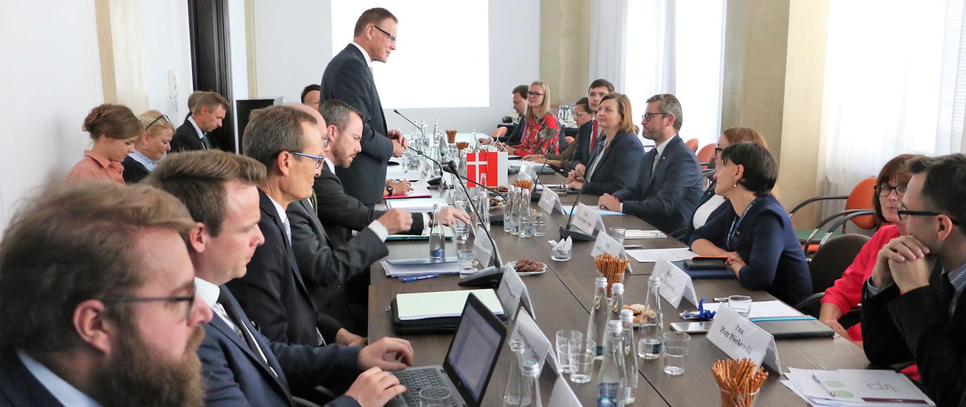 uczestnicy spotkania siedzą wokół stołu . Przemawia przedstawiciel strony duńskiej.