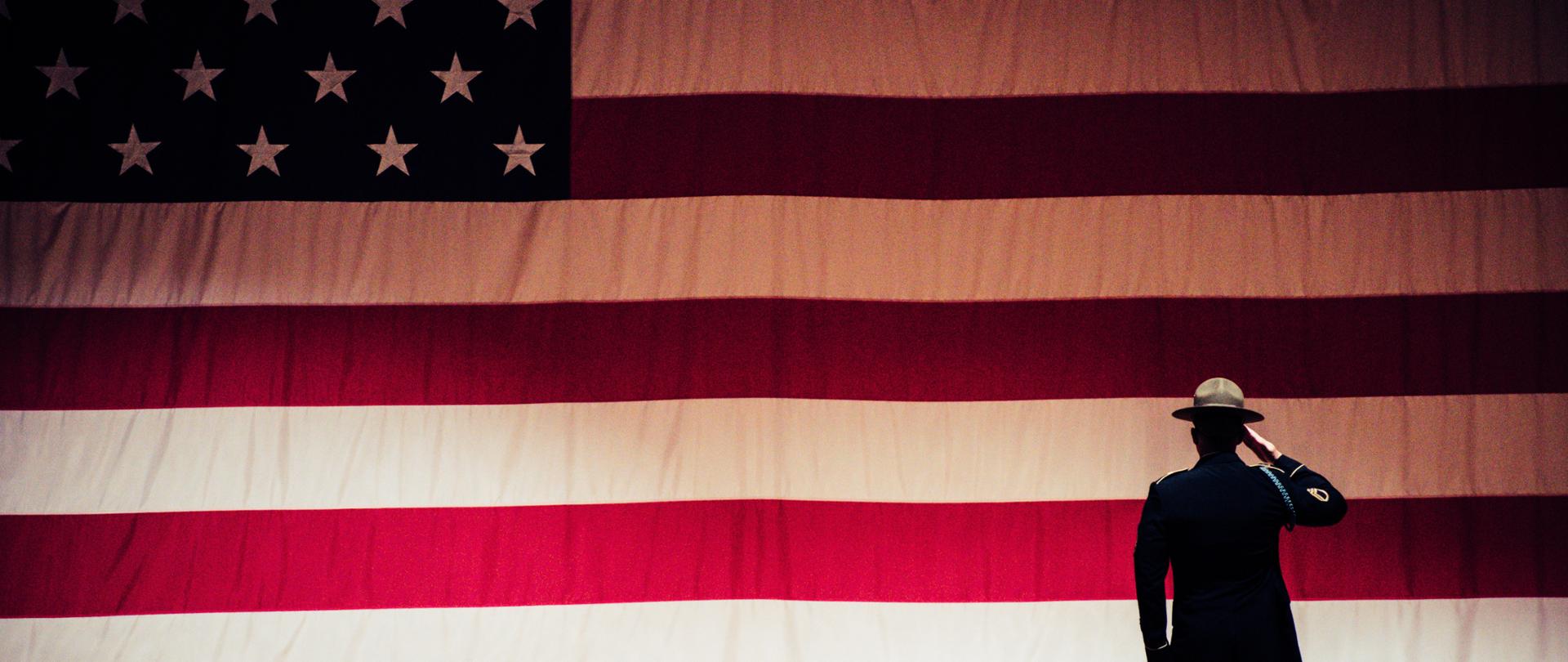 Żołnierz salutujący przed amerykańską flagą.