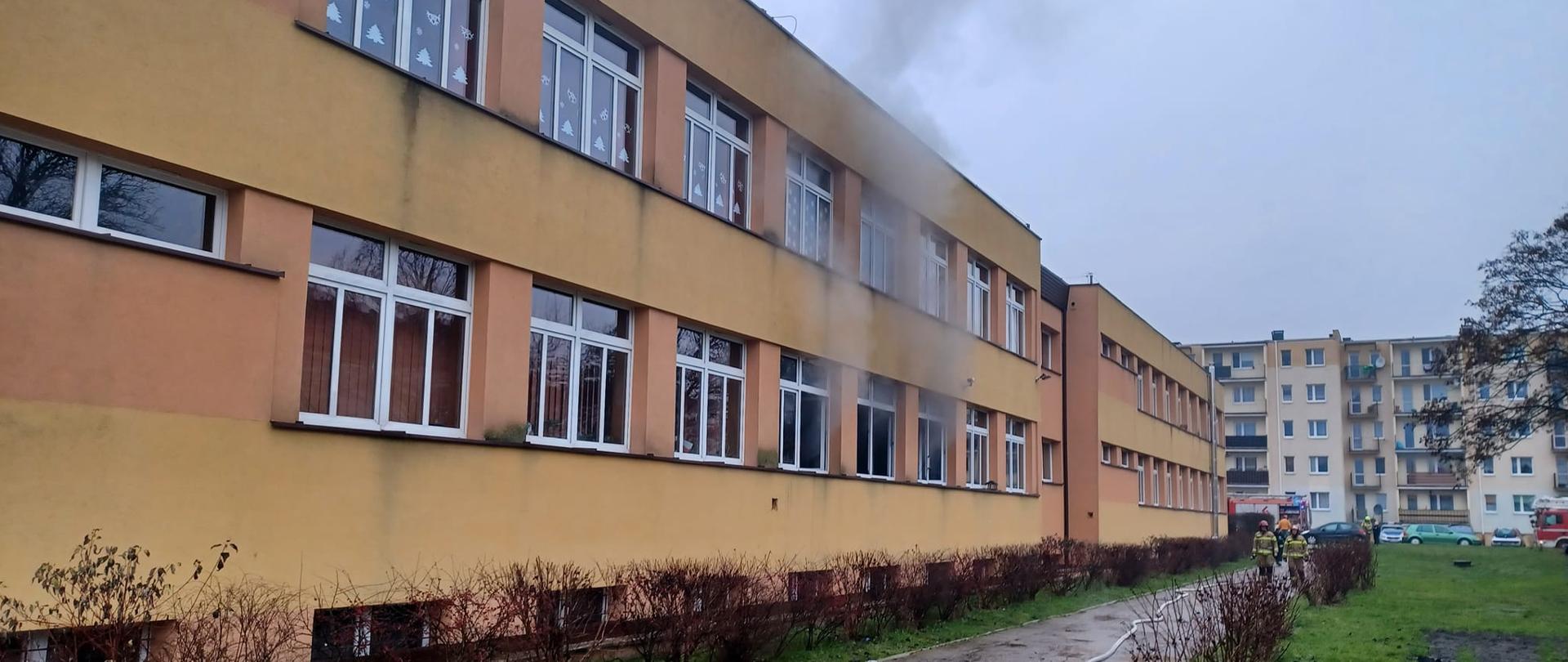 Na zdjęciu widać budynek piętrowy, w którym jest szkoła. Z trzech okien na parterze wydobywa się dym. Przed budynkiem, na chodniku, widać dwóch strażaków w żółtych strojach i czerwonym hełmie oraz rozwinięty biały wąż strażacki. W tle widać samochody strażackie oraz blok. Jest widno.