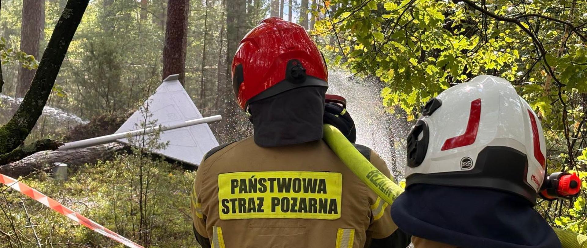 Na pierwszym planie od tyłu widoczni są dwaj strażacy w ubraniach specjalnych i hełmach (czerwonym i jasnym), obserwujący miejsce zdarzenia za biało-czerwoną taśmą ostrzegawczą. Na żółtym pasie na plecach jednego ze strażaków widnieje czarny napis: „PAŃSTWOWA STRAŻ POŻARNA”. W głębi kadru, pośród pni sosen w gęstym lesie, znajduje się wrak białego drona, z którego unosi się biały dym. Pogoda jest słoneczna, a promienie światła malowniczo przenikają przez korony drzew i dym. Zdjęcie wywołuje emocje czujności, gotowości do akcji oraz realnego zagrożenia pożarowego.