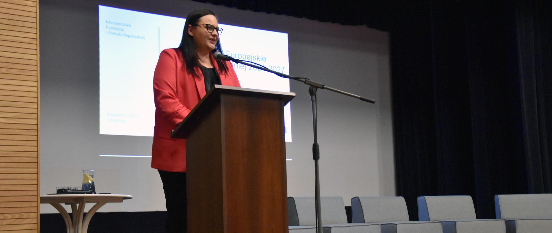 Na scenie w mównicy stoi wiceminister funduszy i polityki regionalnej Małgorzata Jarosińska-Jedynak. Za nią ekran, stolik i w rzędzie fotele.