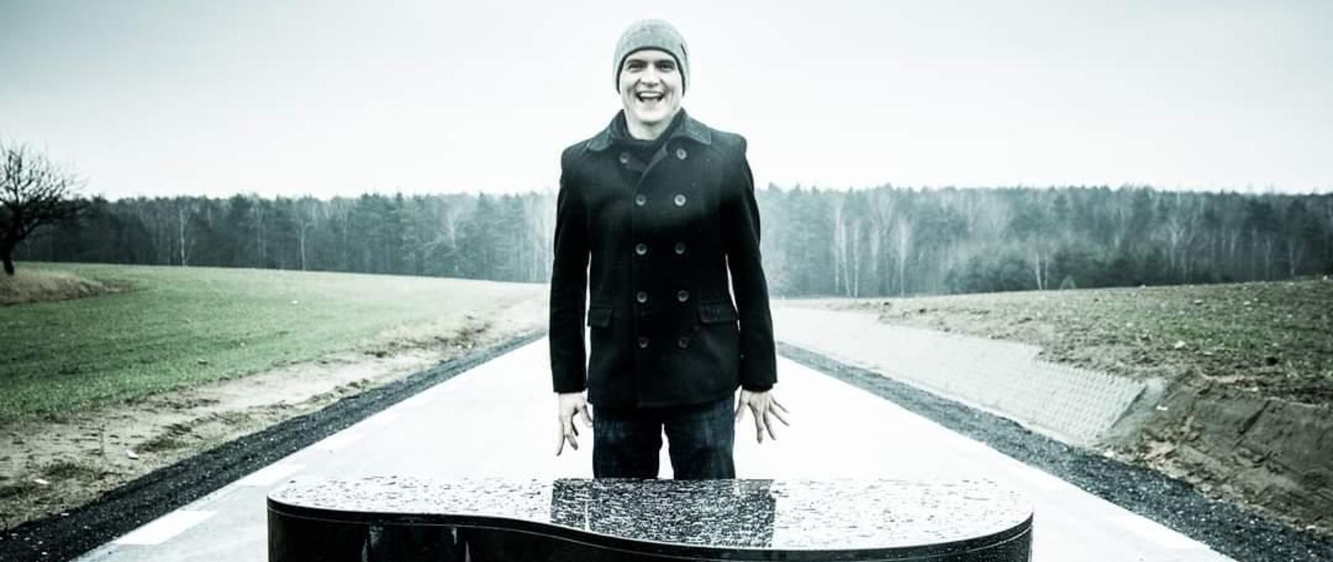 Zdjęcie przedstawia zroszony deszczem czarny fortepian stojący na ulicy pośród pól. Za nim unosi się nad ulicą Maciej Tubis, który ubrany jest w czarny, dwurzędowy krótki trencz i szarą czapkę. W oddali widać las. 