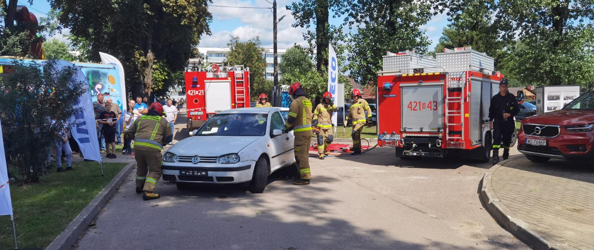Samochód osobowy stojący na drodze przy nim strażacy PSP wykonują pokaz ratownictwa drogowego dla publiczności. W tle pojazdy straży pożarnej oraz publiczność.