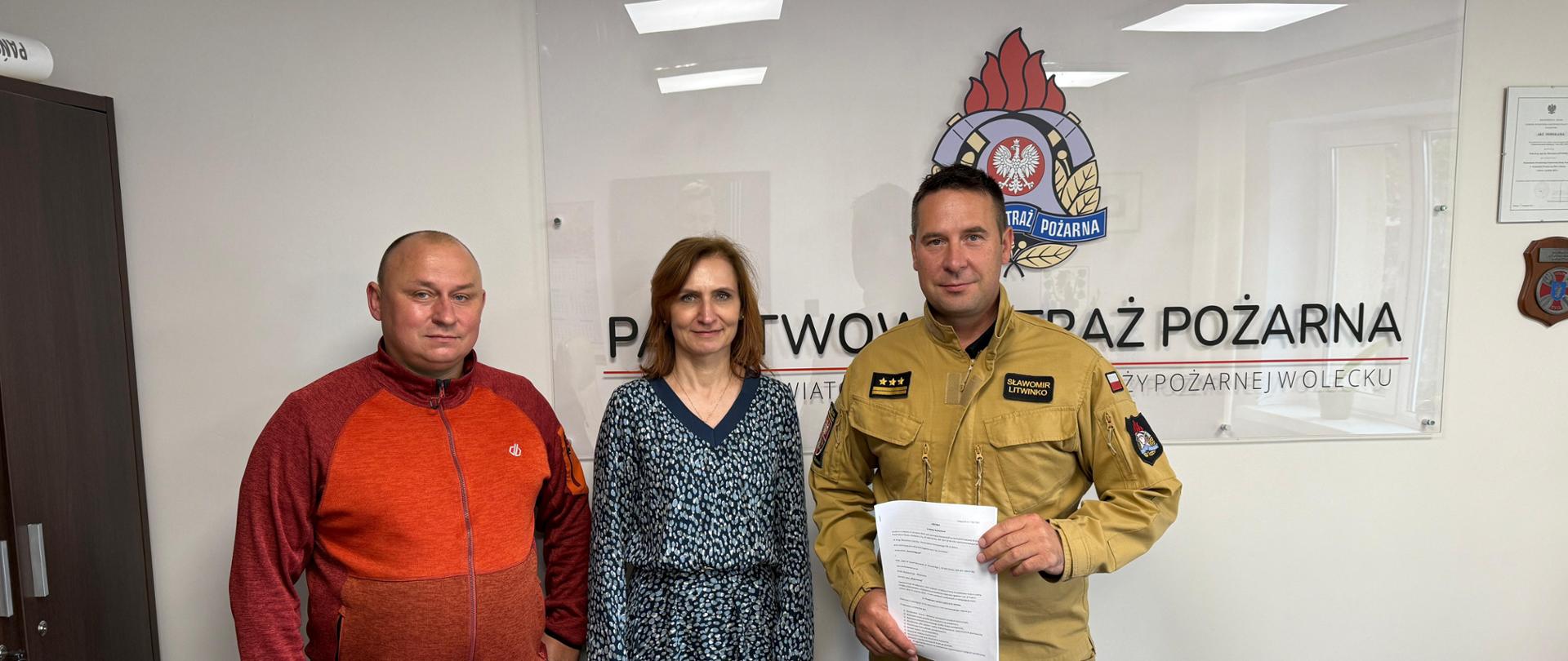 Podpisanie umowy w Komendzie Powiatowej PSP w Olecku – na zdjęciu trzech uczestników wydarzenia stoi przed tablicą z logo Państwowej Straży Pożarnej. Po prawej stronie komendant PSP w mundurze trzymający dokument, obok kobieta w sukience, po lewej mężczyzna w czerwonej bluzie.