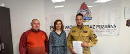 Podpisanie umowy w Komendzie Powiatowej PSP w Olecku – na zdjęciu trzech uczestników wydarzenia stoi przed tablicą z logo Państwowej Straży Pożarnej. Po prawej stronie komendant PSP w mundurze trzymający dokument, obok kobieta w sukience, po lewej mężczyzna w czerwonej bluzie.