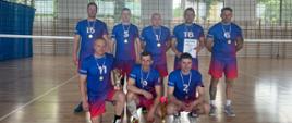 Zdjęcie przedstawia ośmiu mężczyzn w strojach sportowych, pozujących w hali sportowej z widoczną siatką do siatkówki w tle. Mężczyźni ubrani są w niebieskie koszulki i czerwono-niebieskie spodenki. Na koszulkach widoczne są białe numery. Wszyscy mężczyźni mają na szyjach złote medale. Pięciu z nich stoi w tylnym rzędzie, a trzech klęczy w przednim. W rękach trzymają puchar (jeden z nich w przednim rzędzie) oraz dyplom (jeden z nich w tylnym rzędzie). Przed klęczącymi mężczyznami na podłodze stoi puchar.