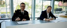 Komendant Główny nadbryg. Wojciech Kruczek oraz Prezes Zarządu PKO Bank Polski Pan Szymon Midera siedzą przy białym stole i podpisują dokumenty. Po lewej stronie komendant Główny PSP w mundurze wyjściowym, patrzy w stronę aparatu. Po prawej stronie Prezes Zarządu PKO Bank Polski w garniturze skupiony na podpisywaniu dokumentu. Za nimi duże przeszklone okna, przez które widać miejską zabudowę i zieleń.