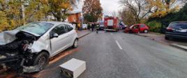 Na zdjęciu widoczny jest srebrny samochód osobowy z uszkodzonym przodem. W tle widoczny jest samochód straży pożarnej, dwie karetki pogotowia oraz czerwony samochód osobowy z uszkodzonym przodem. Widać także pracujących strażaków.
