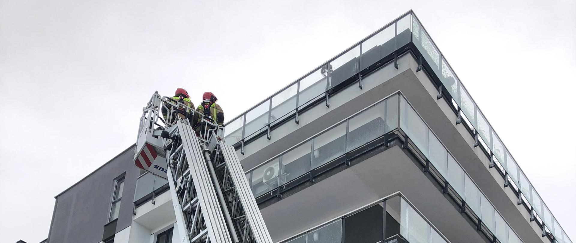 Zdjęcie przedstawia strażaków pracujących na drabinie mechanicznej usuwających szyby w balustradach balkonowych, które zostały uszkodzone przez podmuchy wiatru.