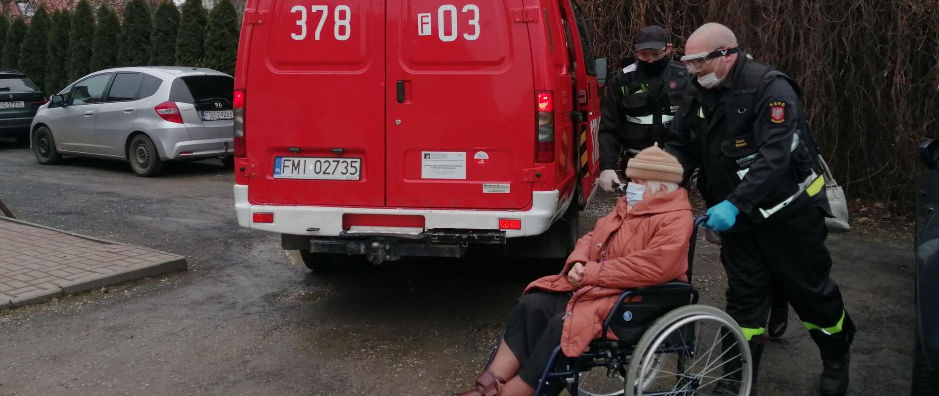 Zdjęcie przedstawia strażaka pchającego wózek inwalidzki z seniorką w miejscowości Lubniewice w dniu 27 stycznia 2021r. W tle czerwony samochód strażacki.