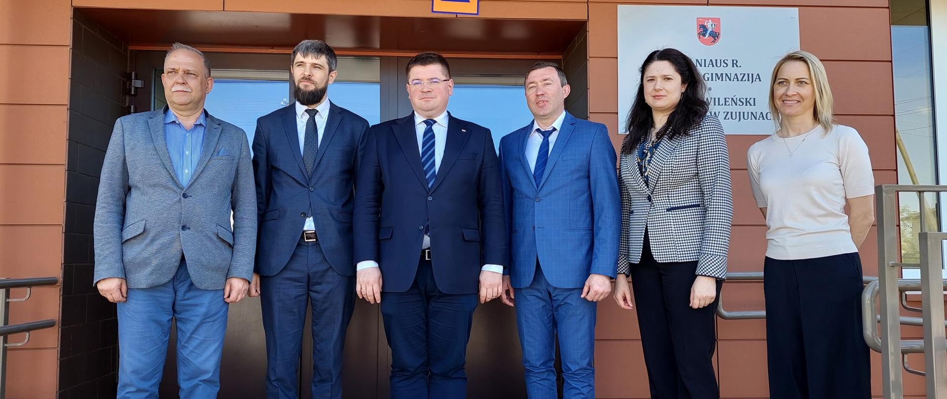 Przed wejściem do brązowego budynku stoi w rzędzie grupa osób, czterech mężczyzn w garniturach i dwie elegancko ubrane kobiety.