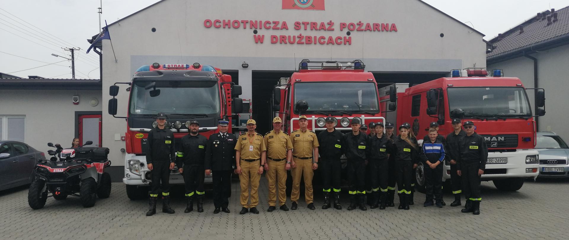 na zdjęciu widzimy strażaków w ubraniach koszarowych na tle samochodów i budynku ochotniczej straży pożarnej