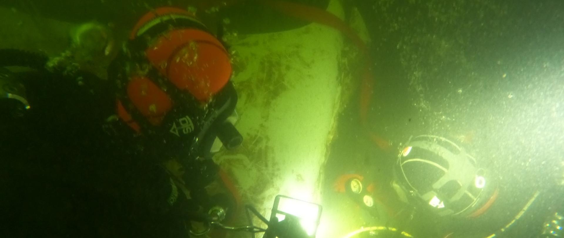 Zdjęcie wykonane pod wodą. Na zdjęciu widać wrak samochodu osobowego. Wrak w kolorze białym. Na wraku dwóch nurków wiąże czerwony pas, który posłuży do wydobycia pojazdu na powierzchnię wody.