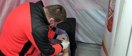 Widok z tyłu. W namiocie – mobilnym punkcie szczepień ratownik medyczny w czerwono-czarnym uniformie pochyla się nad pacjentem i przeprowadza szczepienie. 