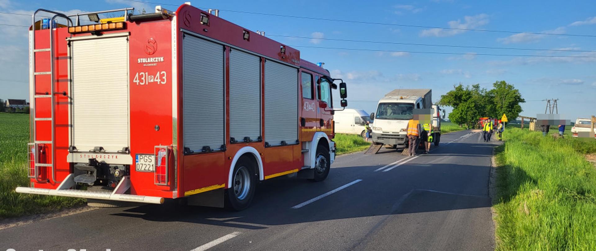 Zdjęcie przedstawia pojazd pożarniczy stojący na drodze. W tle stoi samochód uszkodzony.