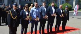 Plac przed komendą. Na czerwonym dywanie stoją przedstawiciele firm i instytuacji wspirających komendę,wraz z komendantem powiatowym i zastępcą. Trzymają w rękach okolicznościowe statuetki. W tle pododdział funkcjonariuszy KP PSP w Rawiczu .