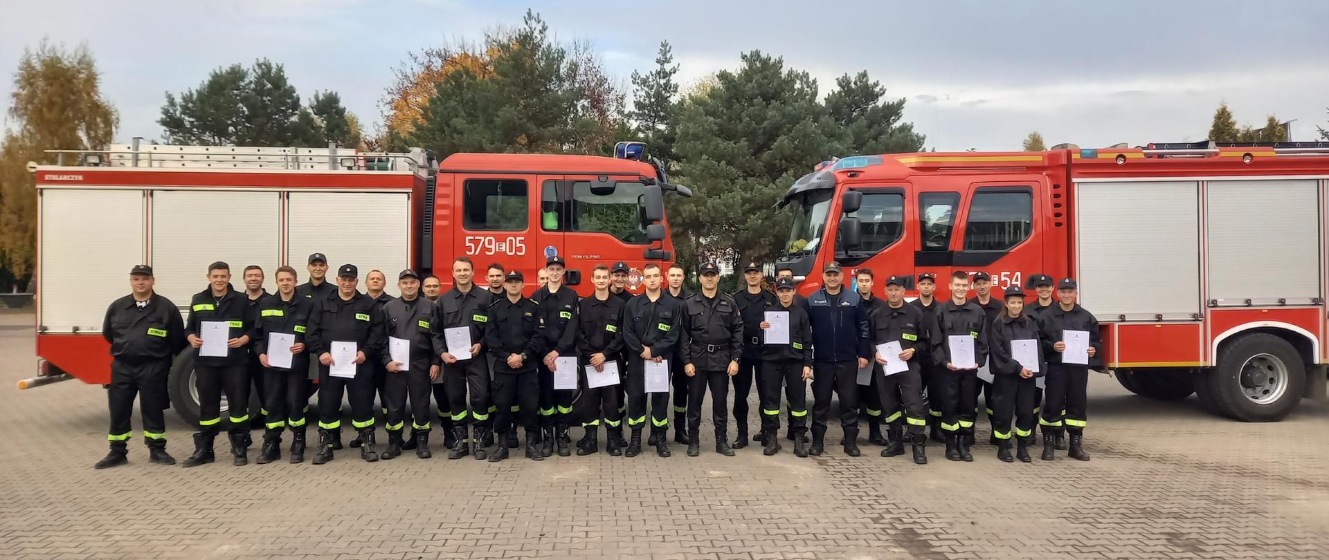 Zdjęcie ilustruje grupę strażaków ratowników OSP w umundurowaniu specjalnym, którzy ukończyli i odebrali zaświadczenia o ukończeniu szkolenia podstawowego. Strażacy stoją w dwuszeregu na tle dwóch pojazdów pożarniczych średnich GBA. Pomiędzy druhami stoją trzej funkcjonariusze KP PSP w Zduńskiej Woli w umundurowaniu koszarowym dowódczo-sztabowym, którzy wręczali zaświadczenia oraz druh Prezes Zarządu Oddziału Powiatowego ZOSP RP w Zduńskiej Woli.