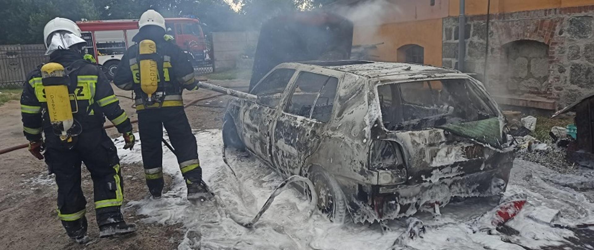 Przedstawia od tył pojazdu marki VW Golf z otwartą maską komory silnika. Strażak w ubraniu specjalnym gasi pojazd pianą gaśniczą