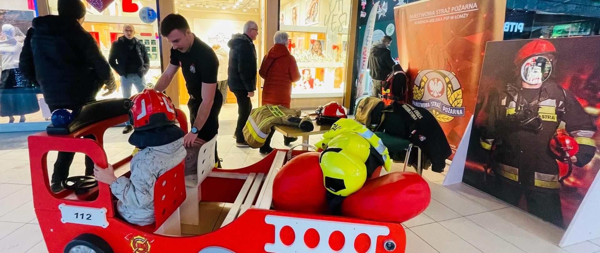 Stoisko przygotowane przez strażaków w Galerii Venda podczas 43 finału WOŚP. Na stoisku znajduję się samochodzik strażacki w którym siedzi mały chłopiec w hełmie, a obok niego stoi strażak. W tle widoczny baner komendy miejskiej PSP w Łomży. 