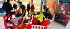 Stoisko przygotowane przez strażaków w Galerii Venda podczas 43 finału WOŚP. Na stosiku znajduję się samochodzik strażacki w którym siedzi mały chłopiec w hełmie, a obok niego stoi strażak. W tle widoczny baner komendy miejskiej PSP w Łomży. 