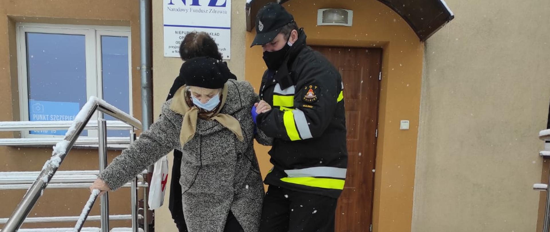 Zdjęcie przedstawia strażaka OSP, który trzyma pod ręką starszą kobietę i pomaga jej zejść ze schodów. W tle znajduje się budynek Niepublicznego Zakładu Opieki Zdrowotnej w Ostrowitem, gdzie znajduje się punkt szczepień. Kobieta oraz strażak mają założone maseczki chirurgiczne na twarzy. Na schodach i poza nimi widać leżący dookoła śnieg. 