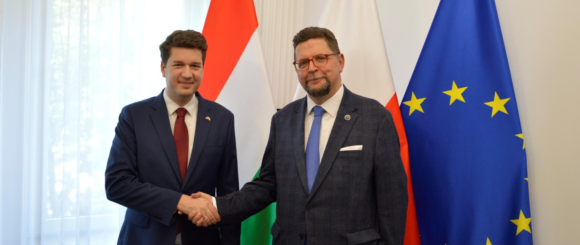Przed białą ścianą stoją wiceminister Szeptycki i mężczyzna w ciemnogranatowym garniturze, podają sobie ręce, za nimi flagi Polski, Węgier i UE, po lewej zasłonięte firankami okno.